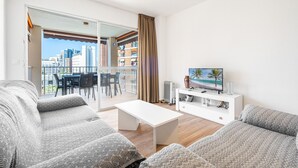 A modern living room with a sofa, coffee table, and TV stand. A sliding glass door leads to a balcony with a city view.
