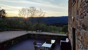 An outdoor patio with stone flooring, a table, and chairs, overlooking a mountainous landscape with trees and a clear sky.