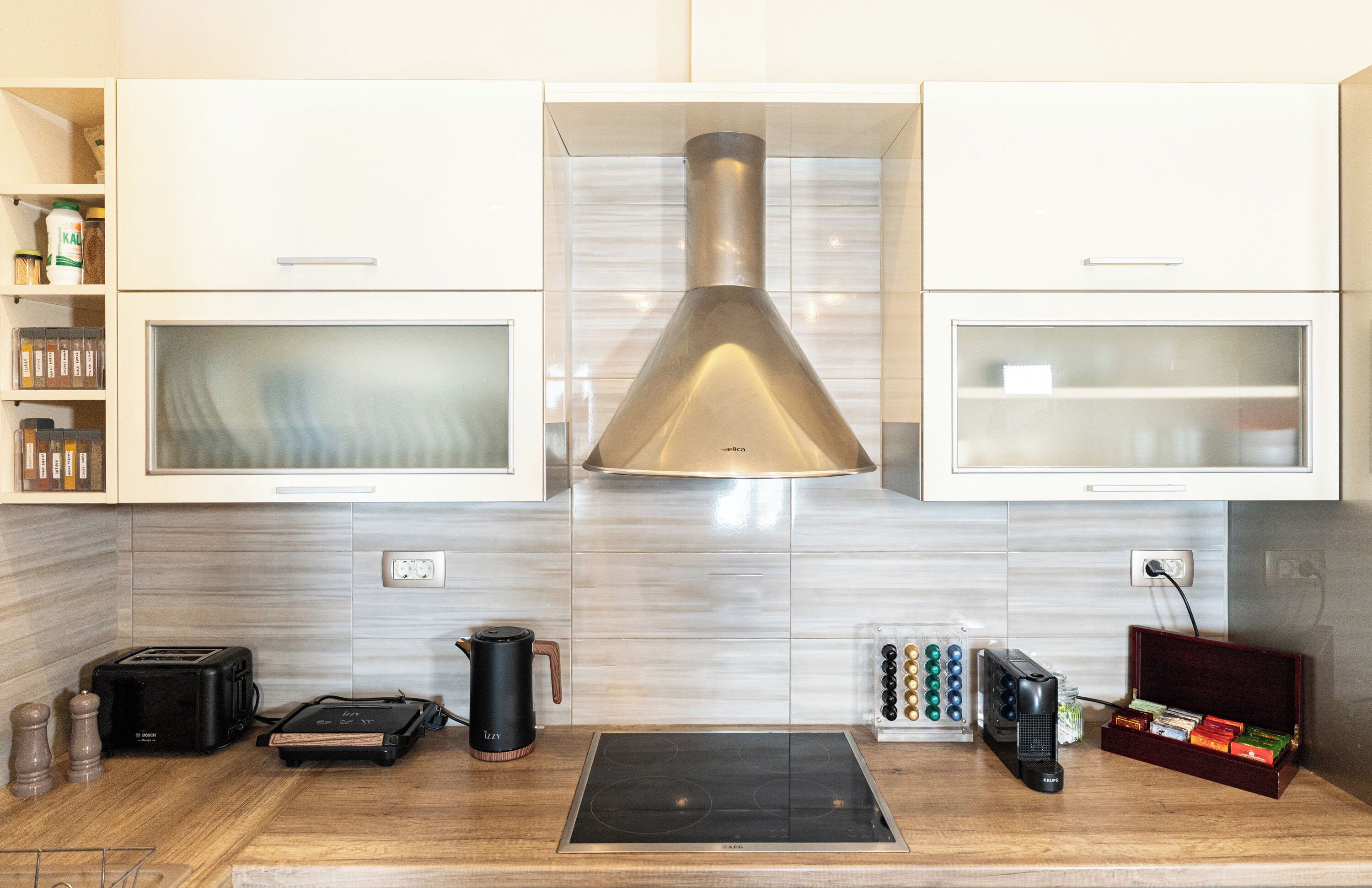 A modern kitchen with white cabinets, a stainless steel range hood, a built-in oven, a coffee maker, a toaster, a kettle, and a small electric grill.