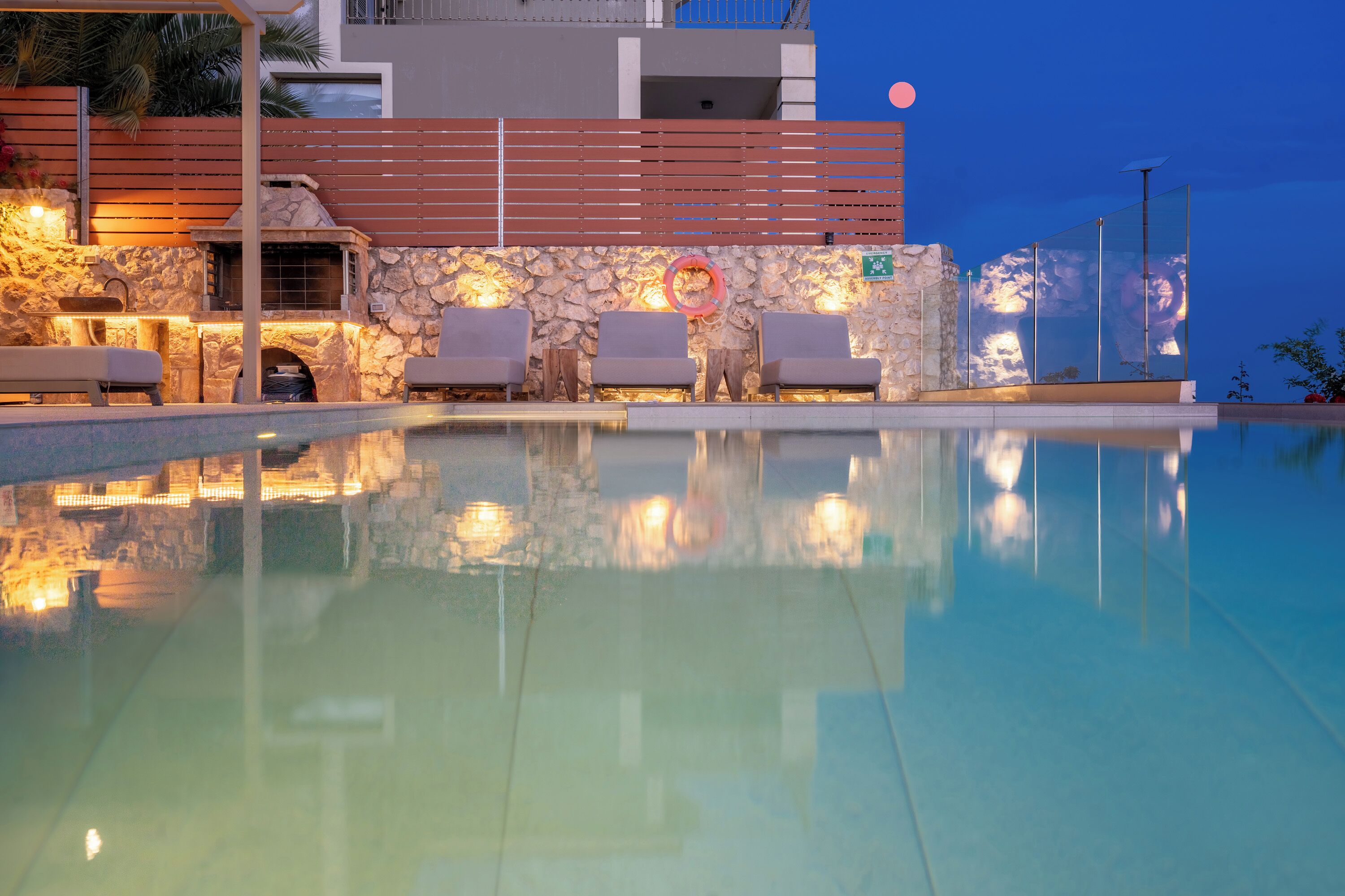 A pool area with lounge chairs, a stone wall, and a sunset in the background.