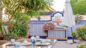 A patio with a glass table set for tea, surrounded by potted plants and a tiled wall with a blue and white pattern.