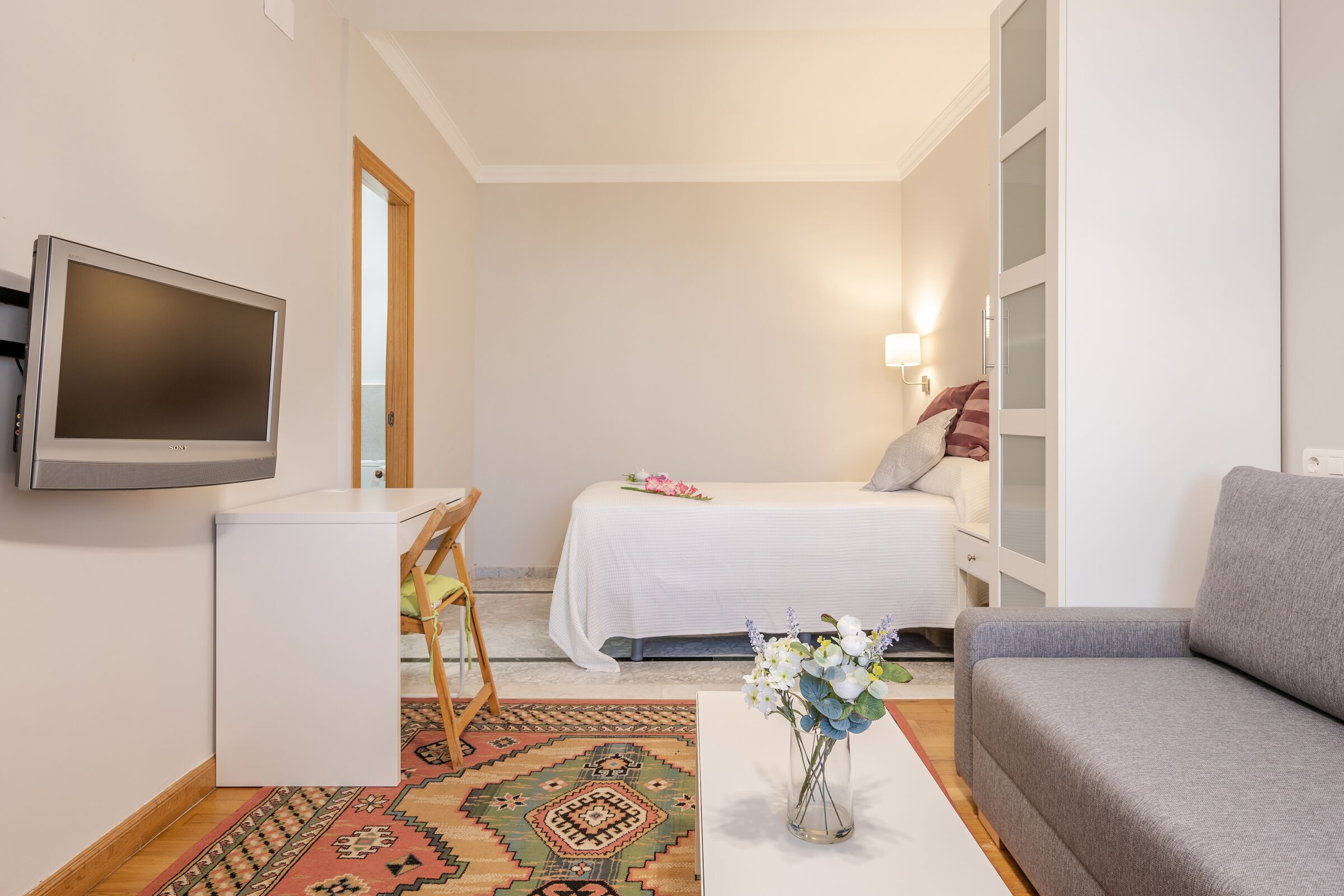 Ein kleines Hotelzimmer mit Bett, Schreibtisch, Fernseher, Sofa und einer dekorativen Vase.