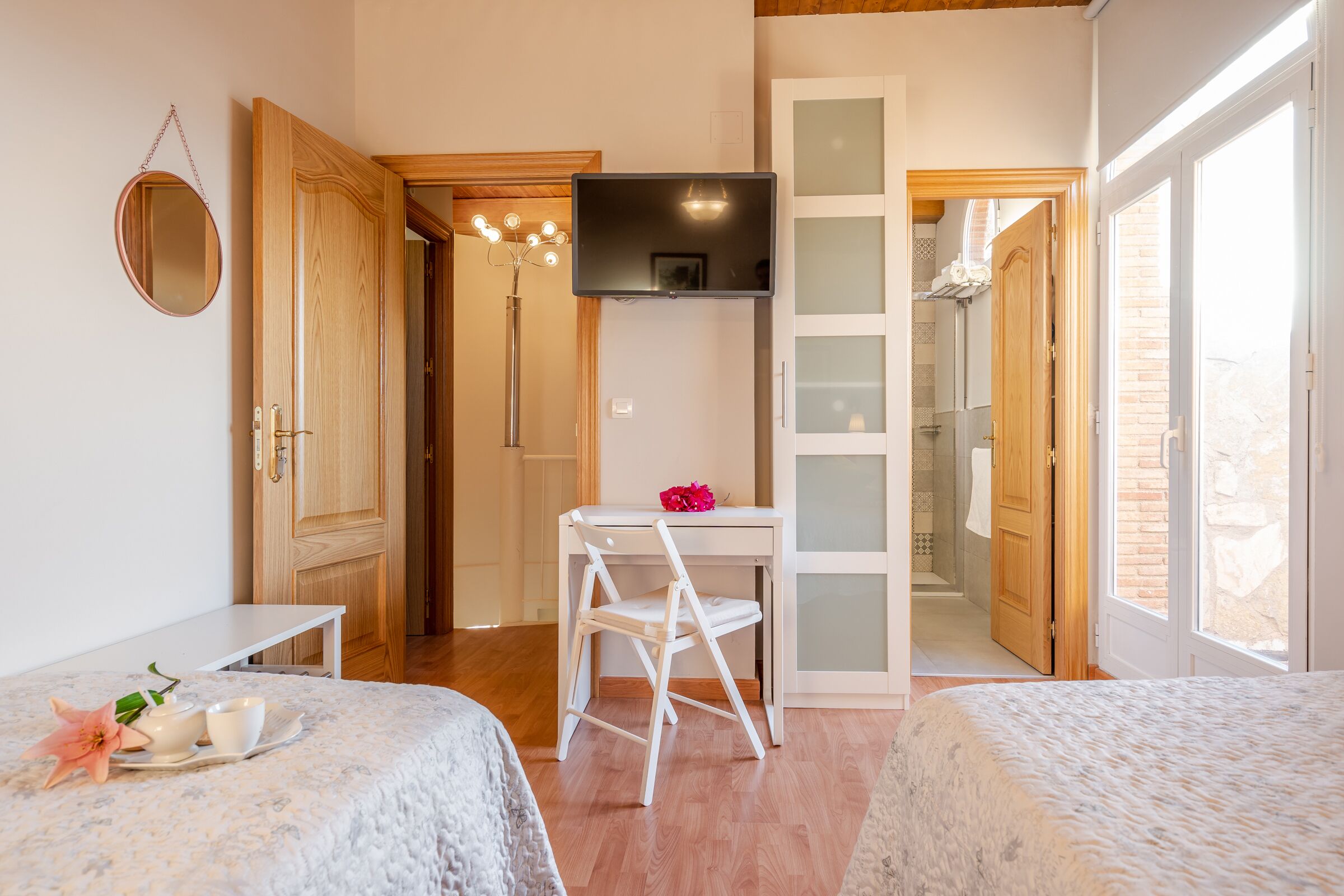 A bedroom with two beds, a desk with a chair, a TV, and a bathroom visible through an open door.
