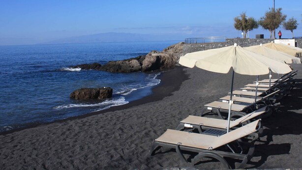 A beach with black sand, a rocky shoreline, and rows of sun loungers with umbrellas. - Farmhouse Finca Delicias La Canaria (Alcalá)