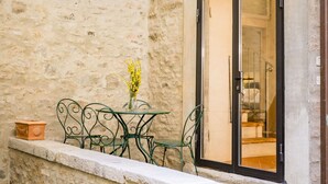 A stone-walled balcony with a table and chairs, a window with a view inside, and a small potted plant. - Appartamento La Corte (Cortona)