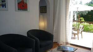 Un salon avec deux fauteuils noirs, une table basse en bois sur laquelle se trouvent un livre et des lunettes, et une porte donnant sur un espace extérieur équipé d’une table et de chaises.