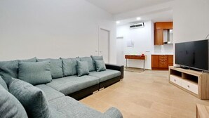 A modern living room with a grey sofa, a TV on a wooden stand, and a kitchen in the background.