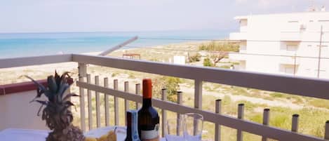 A table with a bottle of wine, glasses, and a fruit bowl on a balcony overlooking a beach.