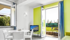 Een moderne kamer met een witte eettafel, witte stoelen, een flatscreen televisie en een glazen schuifdeur naar een buitenruimte met uitzicht op groen.