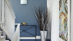 Een gang met een blauwe wand, een witte leuning, een zwart metalen console tafel met decoratieve objecten, en een hoge vaas met takken.