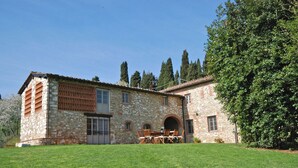 A stone building with a grassy lawn, a patio with chairs, and a large tree.