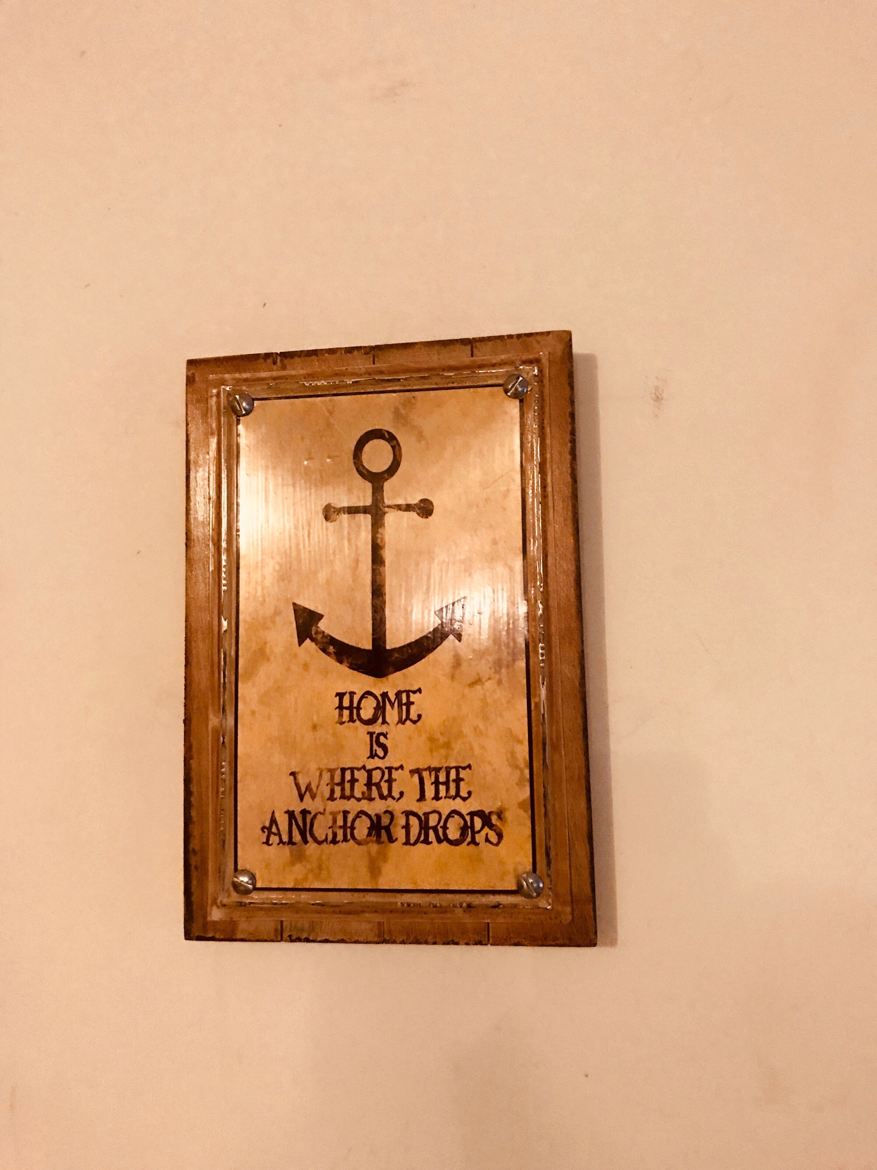 A framed sign with an anchor and the text "HOME IS WHERE THE ANCHOR DROPS" on a plain wall.