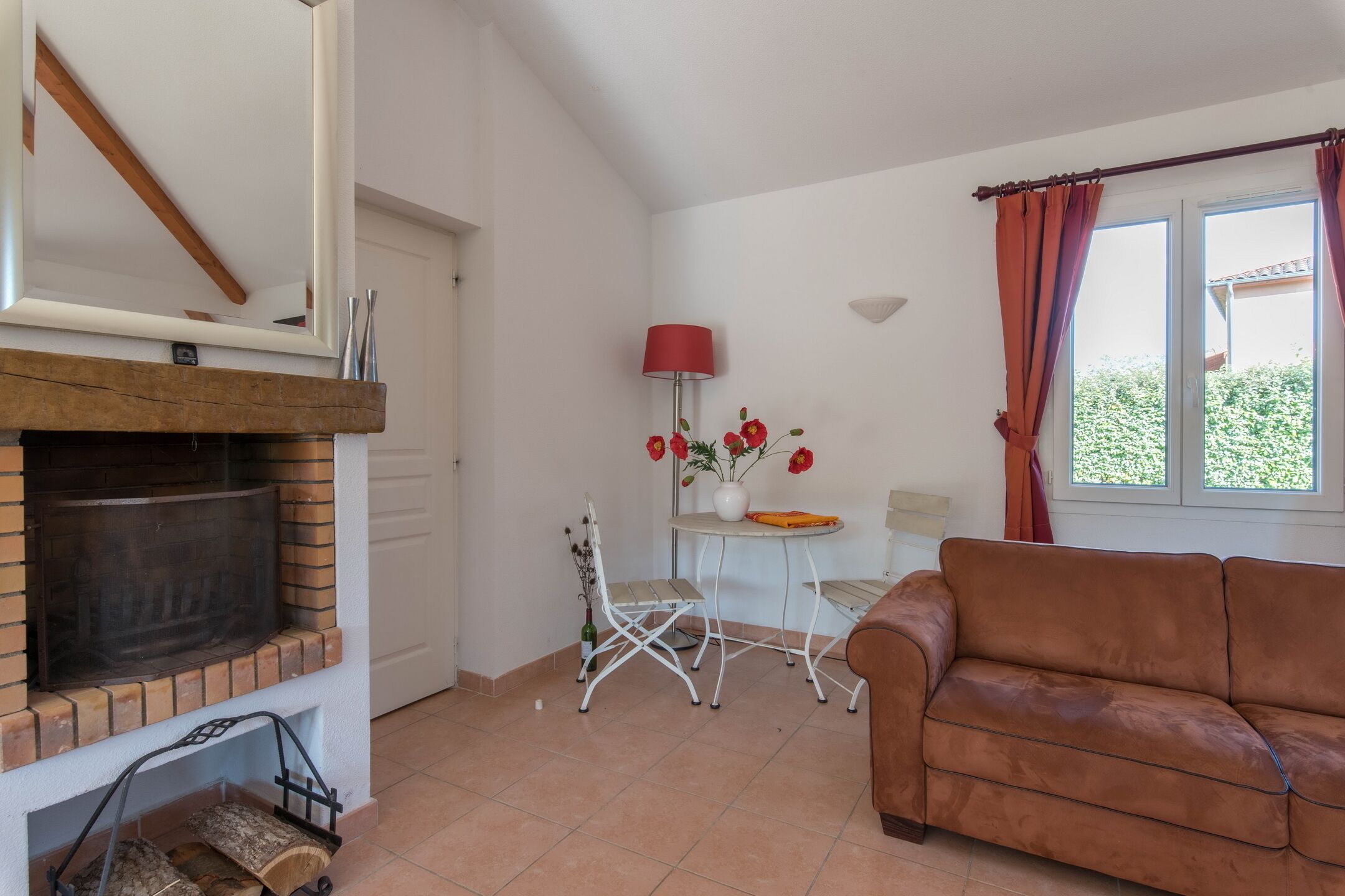 Un salon avec une cheminée, un canapé marron, une petite table avec une lampe rouge et une fenêtre avec des rideaux.