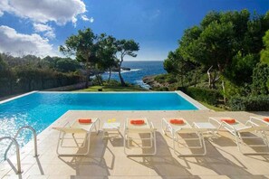 En pool med kristallklart blått vatten, omgiven av solstolar och med utsikt över havet.