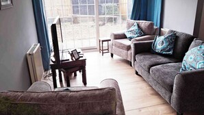 A living room with a grey sofa, a small table, and a chair. There is a window with blue curtains, and a radiator is visible on the wall. - Garden flat, dog friendly, sleeps 3/4, enclosed garden, parking, level access (Tiverton)