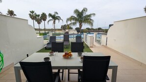 An outdoor patio with a table and chairs, a pool in the background, and palm trees. - 49 - Apartamento Sol Tarifa (Tarifa)