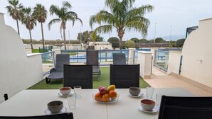 An outdoor dining area with a table set for two, surrounded by chairs, with a view of a pool and palm trees in the background. - 49 - Apartamento Sol Tarifa (Tarifa)