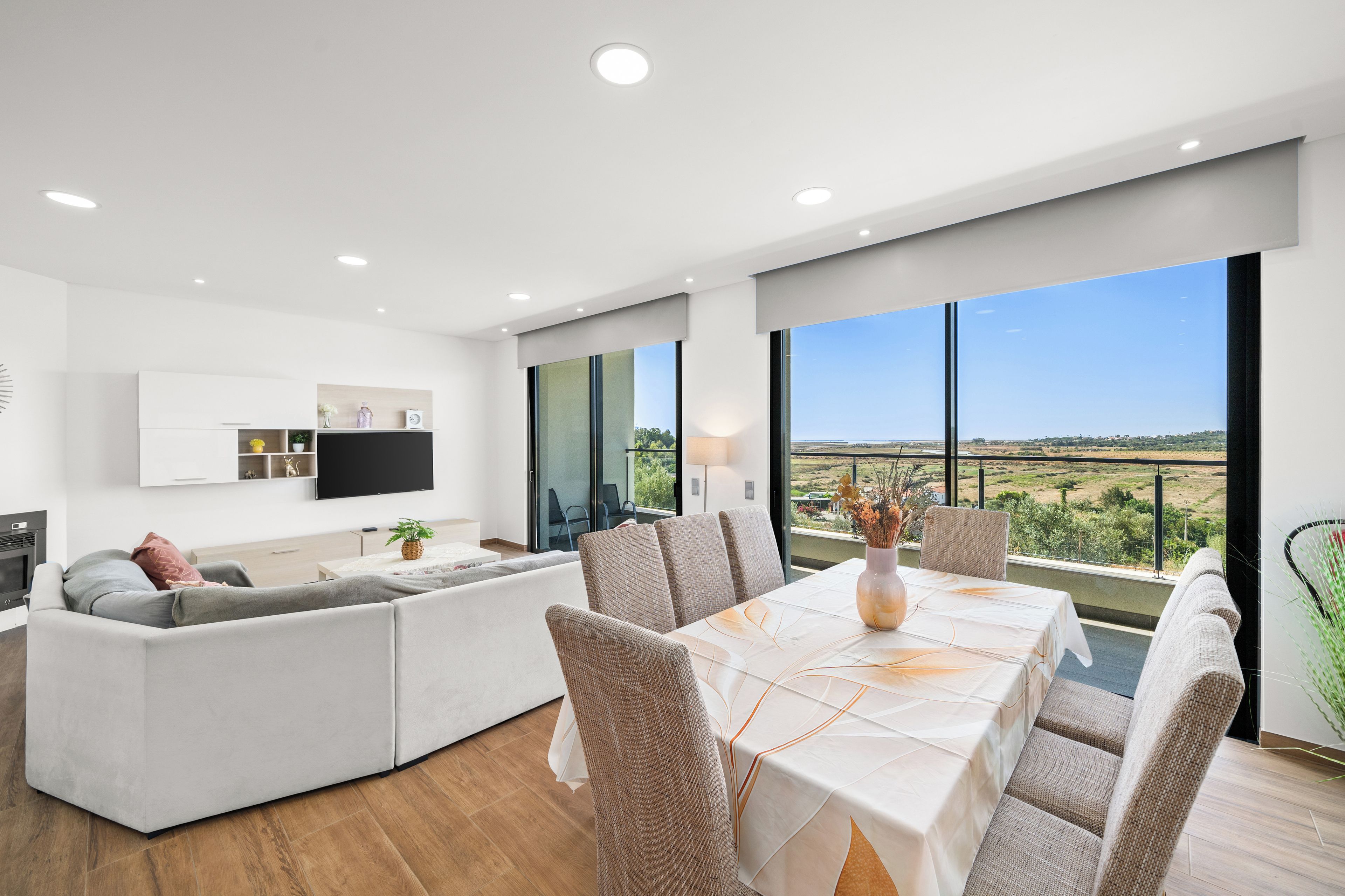 A modern living room with a sofa, dining table, and chairs, a flat-screen TV, and a large window with a view of the countryside.
