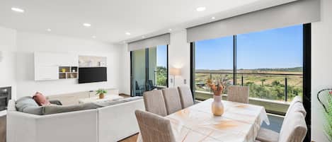 A modern living room with a sofa, dining table, and chairs, a flat-screen TV, and a large window with a view of the countryside.