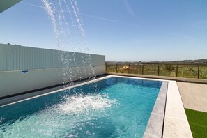 A rectangular swimming pool with clear blue water, surrounded by a concrete deck and a metal railing.