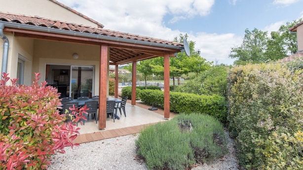 Une terrasse couverte, aménagée avec du mobilier d’extérieur, entourée d’un jardin bien entretenu et d’une maison dont le toit est recouvert de tuiles.
