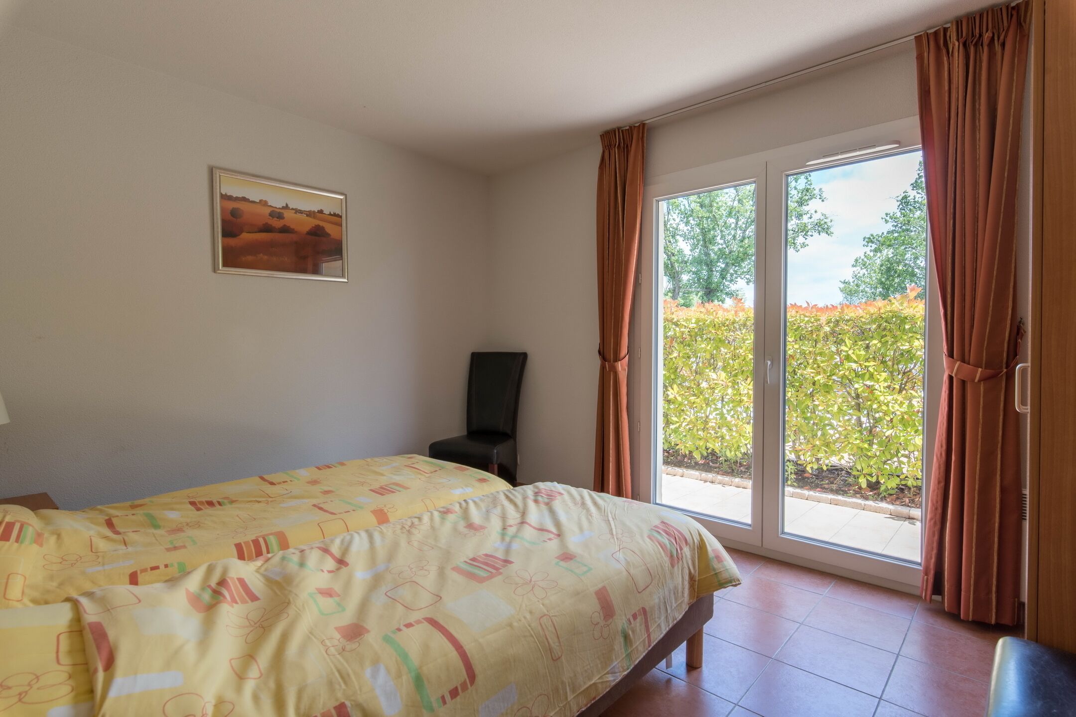 A bedroom with a bed, a chair, a window with curtains, and a painting on the wall.