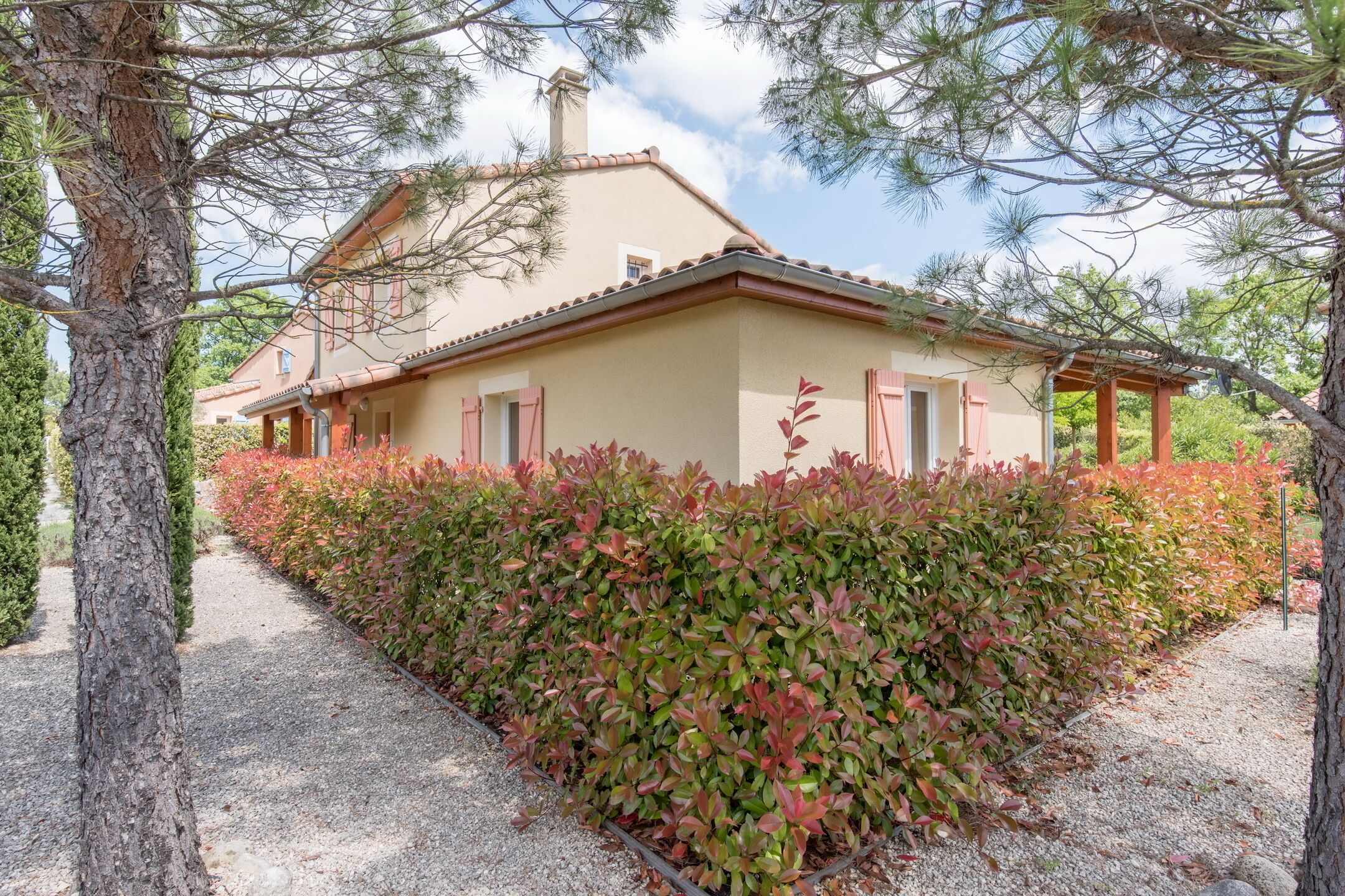 A house with a beige facade, red shutters, and a tiled roof, surrounded by a gravel path and dense shrubbery.