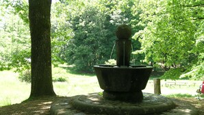 Une fontaine en pierre surmontée d’une sphère, entourée d’une base circulaire également en pierre, située dans un parc arboré avec une pelouse.