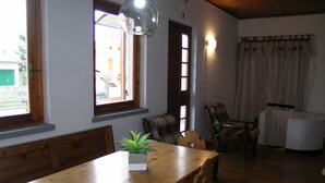 Un coin repas avec une table en bois, des chaises et une plante en pot. Il y a des fenêtres avec des rideaux et une lampe suspendue au-dessus de la table.