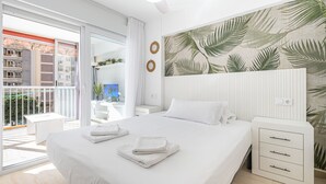 A modern hotel room with a large bed, a balcony with a view of buildings, and a tropical leaf-patterned wall. - Carabelas III 1-3 (Benidorm)