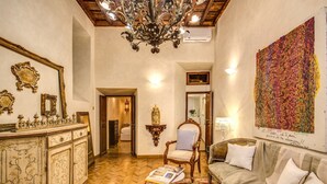 A spacious room with a wooden floor, a large chandelier, a sofa, and a wall with a colorful abstract painting. - 1 Min to Pantheon – Designer 2BR Apt with 3 AC + Elevator (Roma)