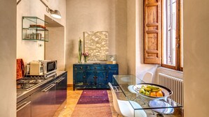A compact kitchen with a gas stove, microwave, and a blue cabinet. There's a glass table with a fruit bowl and a vase with flowers. - 1 Min to Pantheon – Designer 2BR Apt with 3 AC + Elevator (Roma)