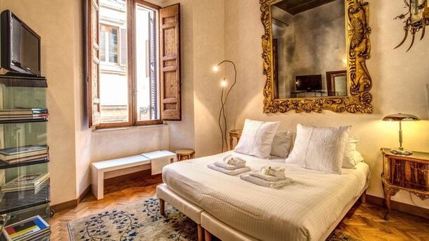 A bedroom with a large bed, a wooden bench, a bedside table, a mirror, and a bookshelf. - 1 Min to Pantheon – Designer 2BR Apt with 3 AC + Elevator (Roma)