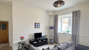 A modern living room with a grey sofa, a black entertainment unit, a small table with a vase of flowers, and a window with curtains. - Central Apartment Linlithgow, near Edinburgh (Linlithgow)