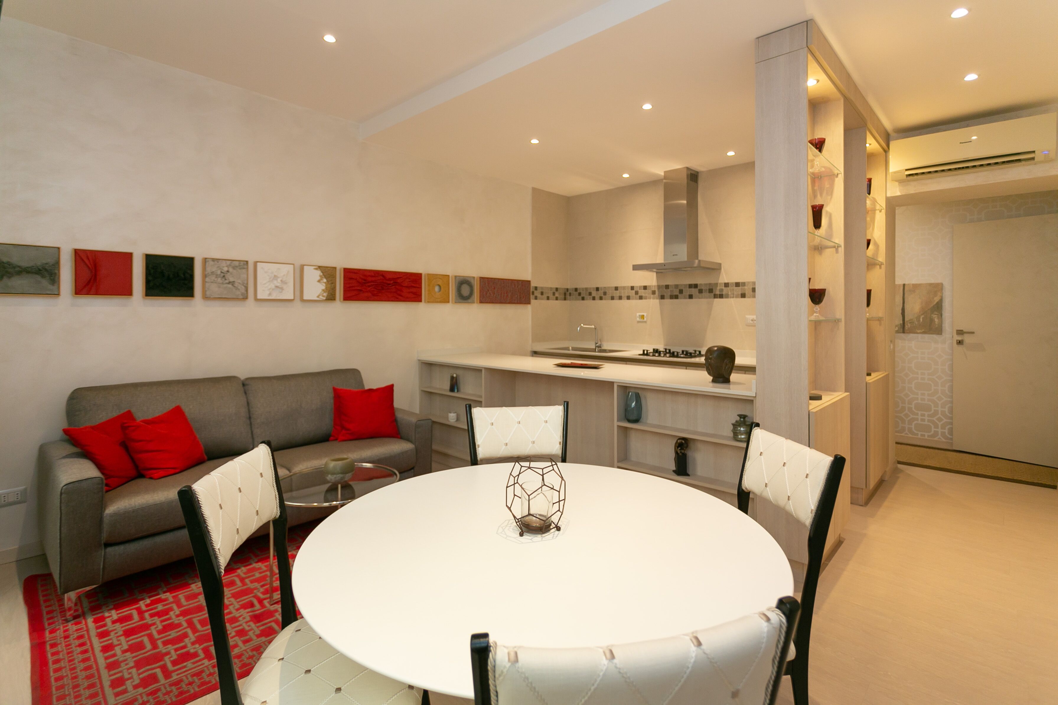 A modern living room with a round dining table, grey sofa with red cushions, and a kitchen in the background.