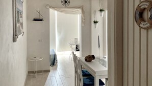 A narrow hallway with a white door on the right, a white wall with a framed picture, and a small white table with a chair.