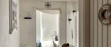 A narrow hallway with a white door on the right, a white wall with a framed picture, and a small white table with a chair.