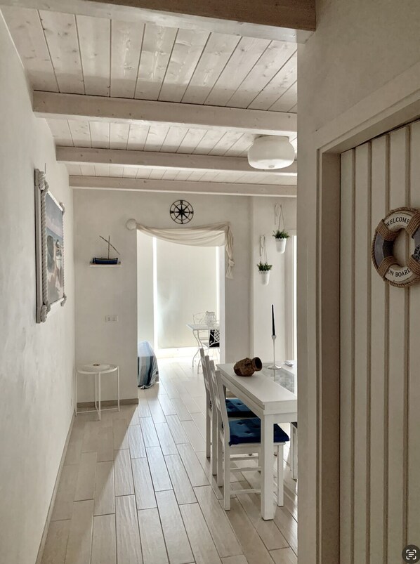 A narrow hallway with a white door on the right, a white wall with a framed picture, and a small white table with a chair.