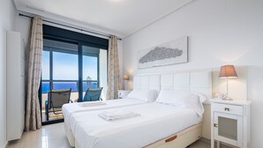 Habitación de hotel con dos camas, un balcón con una silla, cabecera blanca, mesita de noche con lámpara y vistas a la ciudad.