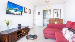 Una sala de estar con un sofá rojo, un televisor montado en la pared, un mueble de entretenimiento de madera y una mesita auxiliar con mandos a distancia.