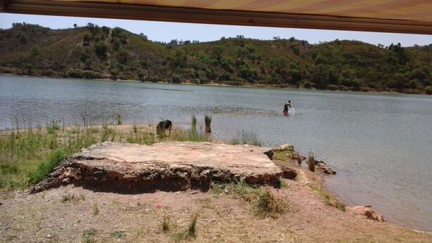 Paisaje natural con una masa de agua, un afloramiento rocoso en primer plano y un terreno montañoso al fondo.