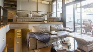 Un espace de vie moderne de style loft, comprenant un canapé, une table basse en verre et une vue sur l’extérieur grâce à de grandes fenêtres.