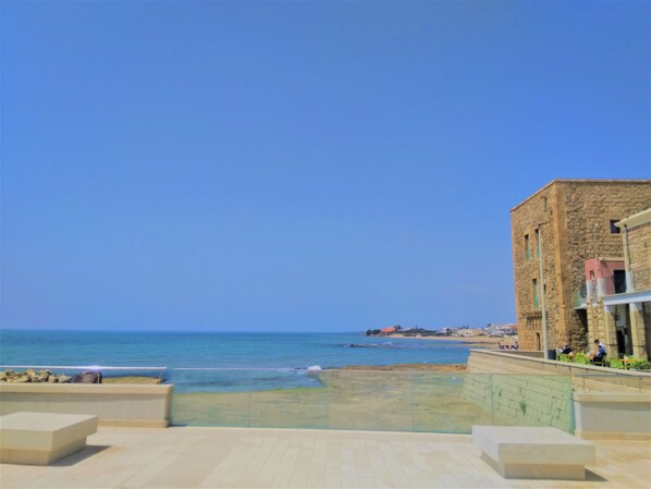 Vistes costaneres amb un cel serè, una platja de sorra i un edifici de pedra amb un balcó.