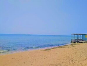 Une plage avec une eau d’un bleu limpide, un rivage sablonneux et un petit bâtiment surmonté d’un toit bleu.
