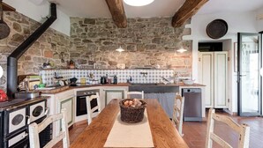 Una cocina rústica con pared de piedra, vigas de madera, una larga mesa de madera y una estufa.