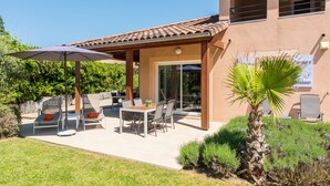 Une terrasse couverte, aménagée avec du mobilier d’extérieur, un palmier et une vue sur la maison.