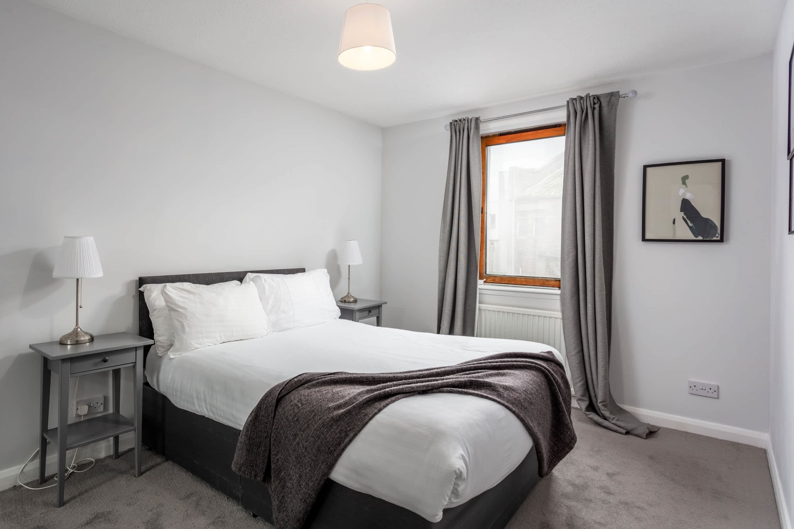 A neatly arranged bedroom with a bed, bedside lamps, a nightstand, a window with curtains, and a framed picture on the wall.