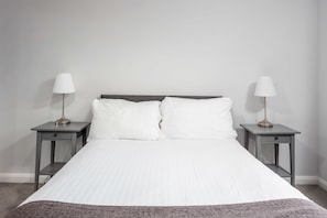 Cama com roupa de cama branca, duas mesas de cabeceira com abajures e uma parede lisa.
