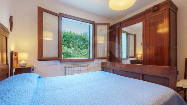 Une chambre avec un grand lit, une fenêtre donnant sur un paysage verdoyant, une armoire en bois et une table de chevet avec une lampe.