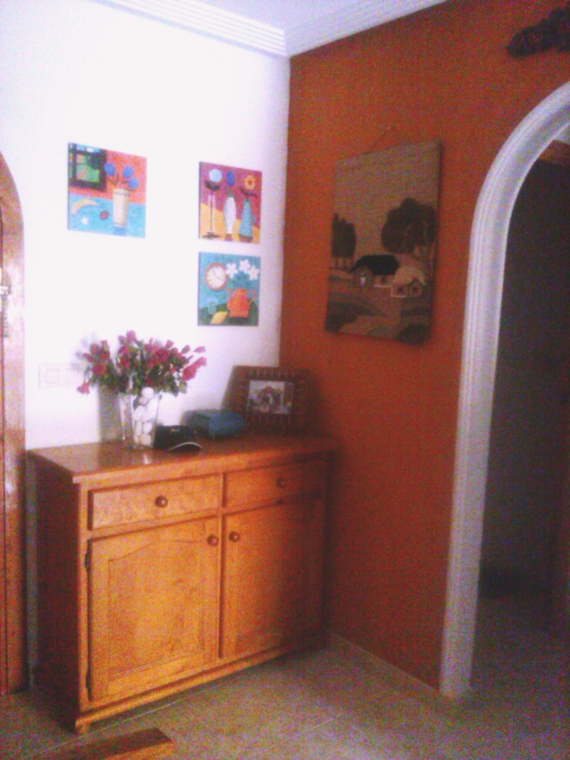 A room with a wooden cabinet, a vase with flowers, and three paintings on the wall.